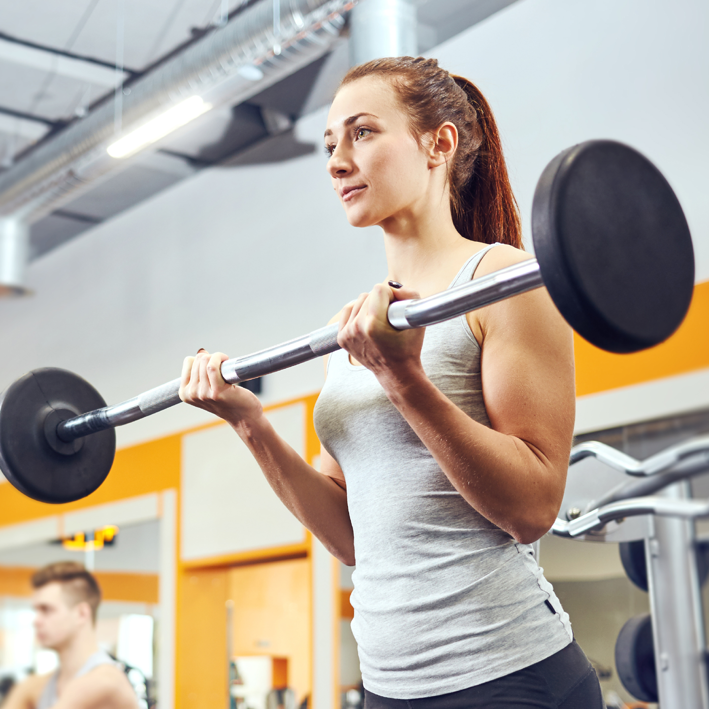 A Caucasian female in her twenties is captured lifting a bar bell. She is wearing a grey tank top and her hair is in a high pony tail. There is a Caucasian male captured to the left of the frame also working out in the gym at the YMCA.