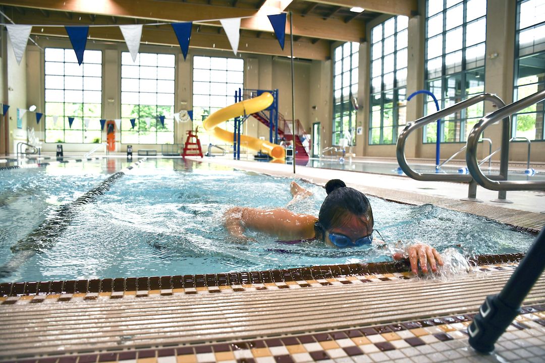 An image of a young Asian female practicing for the swim team at the YMCA's pool. She is wearing goggles and is captured in the middle of a butterfly stroke. Her hair is pulled into a bun, and her nose and mouth and down in the water.