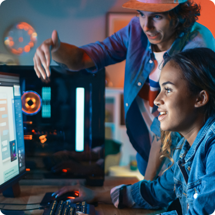 African American female teen playing Esports on a computer. With a caucasian teen male standing next to the female coaching her how to play. 