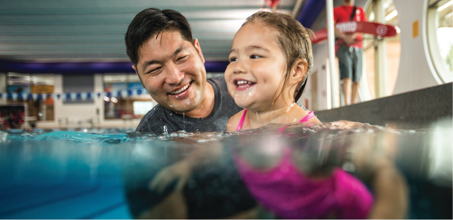 Father and daughter swim in a ymca indoor pool and smile spending quality time together