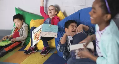 An image taken in a classroom at one of the YMCA's of four children laughing during reading time. From left to right, the Asian boy, Caucasian girl, Hispanic boy, and African American girl are all sitting on bean bags and holding a book in their hands.