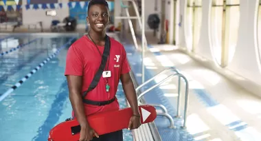 An image of an African American male lifeguard wearing a red YMCA branded t-shirt, and red buoy life-saving device. He is captured standing and smiling in front of the indoor YMCA pool.
