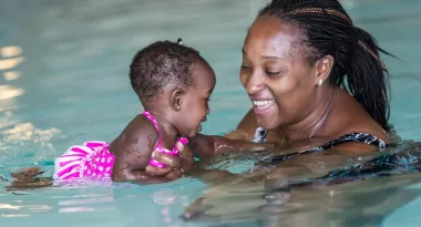 An image of an African American mother and her infant daughter partaking in swim lessons at the YMCA. Both are captured smiling at one another.