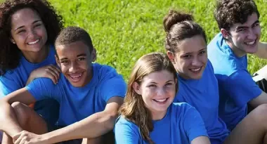 A diverse group of five YMCA volunteers sitting on the ground, wearing matching blue t-shirts. There are three females and two males in the group.