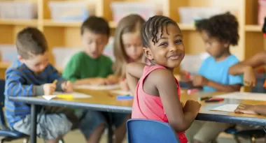 A young African American girl poses looking back towards the camera with a soft smile on her face as she is sitting, coloring pieces of papers with colorful markers with her diverse classmates at a table in the room at the YMCA.