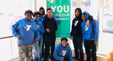 An image of a group of African American teens from the YMCA teenage program posing for a photo in front of a YMCA sign, wearing YMCA branded black and blue colored hoodies. 