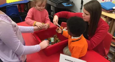 An image of a Caucasian female staff member at the YMCA helping three after school children, two Caucasian girls and one African American boy, ranging in ages. They are all playing with a box filled with small figurines.