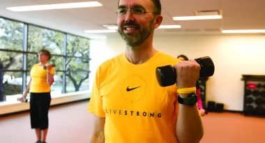 An image of a Caucasian male lifting a weight with his left arm during a fitness class at the YMCA. He is wearing a yellow shirt with black writing on it that says "Livestrong" and he is wearing glasses and has a focused expression on his face.