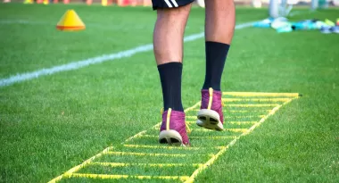 An image of a pair of men's legs wearing purple tennis shoes, black socks, and black shorts whilst doing a ladder drill on a grass field at the YMCA preparing for a soccer game. 