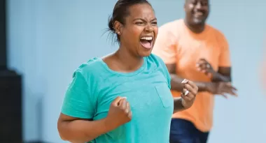 An image of an African American woman posing triumphantly with her fists closed, like she is excited to workout. An African American male is captured smiling at her in the background. On the right side of the frame, a Caucasian female is barely visible.
