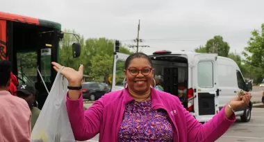 An image of an African American woman posing with a bag full of goodies outside of a YMCA. She is wearing a pink jacket, purple shirt with a swirling pattern, and silver, hoop earrings.