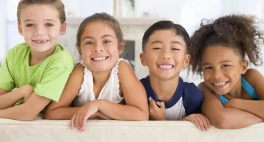 An image from left to right of two Caucasian children, a boy and a girl, an Asian boy, and an African American girl crossing their arms on the back of the couch, smiling for the camera.