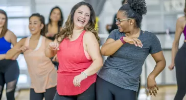 An image of a Caucasian female and African American female both bumping into one another's sides as they participate in a dance class with a diverse group of women at the YMCA.