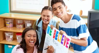 An image of three teens making a colorful sign together that says, "we believe," as well as other words like "respect." The African American female, Asian female, and Middle Eastern male are all looking at the camera, smiling.