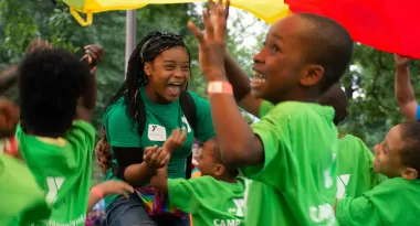 A female, African American camp leader at the YMCA leads the children in excitement for a day at YMCA Camp. The male and female African American children are all dressed in the same lime green YMCA t-shirts and are throwing a parachute up above their heads.