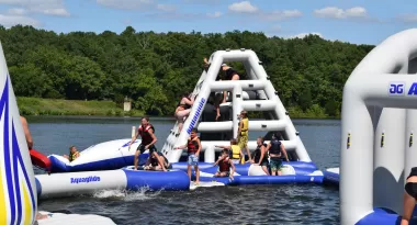 An image of a group of young girls and boys wearing life vests and playing on an inflatable water park placed on a lake at the YMCA.