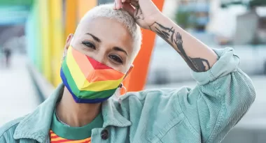 An image of an African American girl with a shaved head that has been dyed blonde, is captured wearing a rainbow pride mask. She is also wearing a blue button up shirt, and a red and orange striped shirt with a green collar. Her left arm is resting on her head, making the tattoos on her forearm visible.