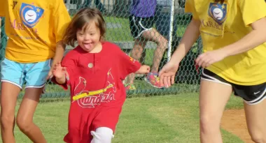 A happy young girl with Down Syndrome in the Miracle League baseball program runs to first base in a St. Louis Cardinals team jersey.