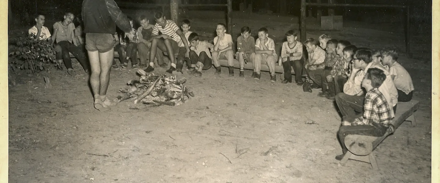 A group of young boys, original Camp Lakewood campers from the 1950s, sitting around a roaring campfire at night.