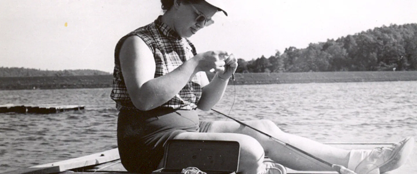A woman in 1960s attire smiles while carefully baiting a fishing hook by the shore of Sunnen Lake.