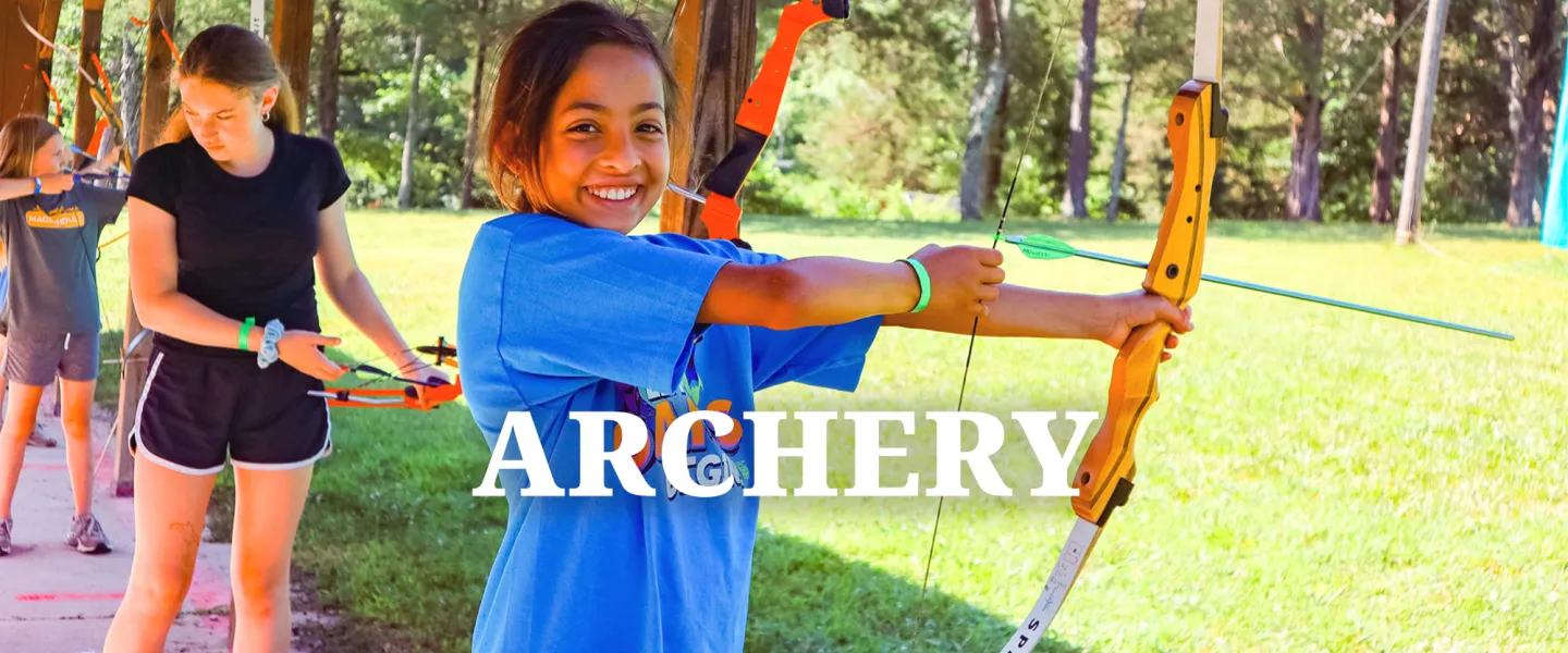 A camper smiling while aiming a bow and arrow at a target on the archery range.