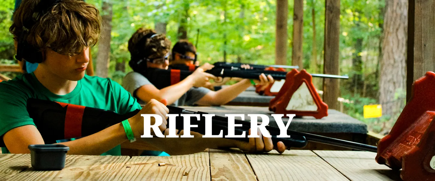 A camper wearing eye and ear protection aiming an air rifle at a target under supervision.