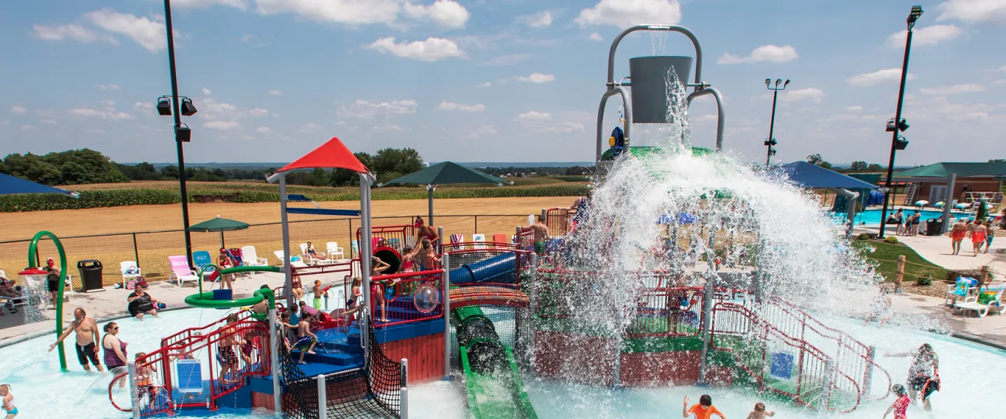 Children playing on the colorful multi-level splash pad structure at River Rapids Waterpark, featuring small slides and interactive water sprays under a bright blue sky.