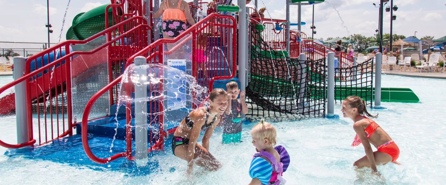 Young toddlers playing safely in the shallow, zero-depth entry splash pad area featuring small, colorful slides and interactive water sprayers.