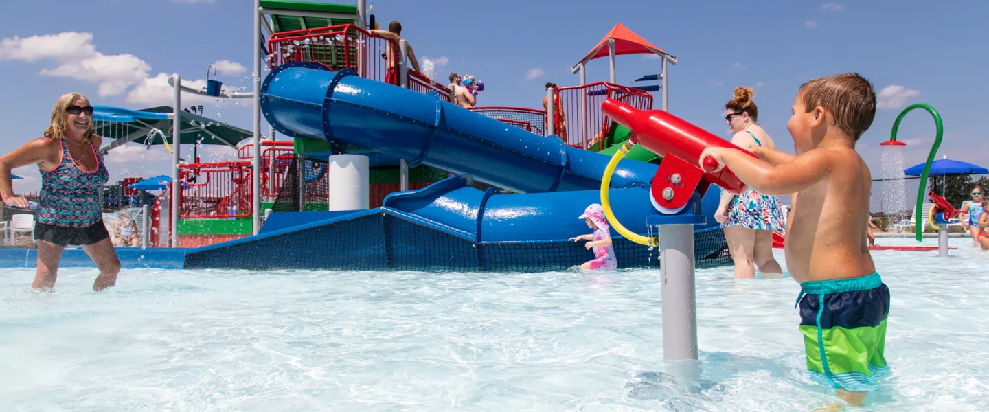 A young boy in the shallow pool interacting with a red water shooter station at the Ste. Genevieve YMCA River Rapids Waterpark.