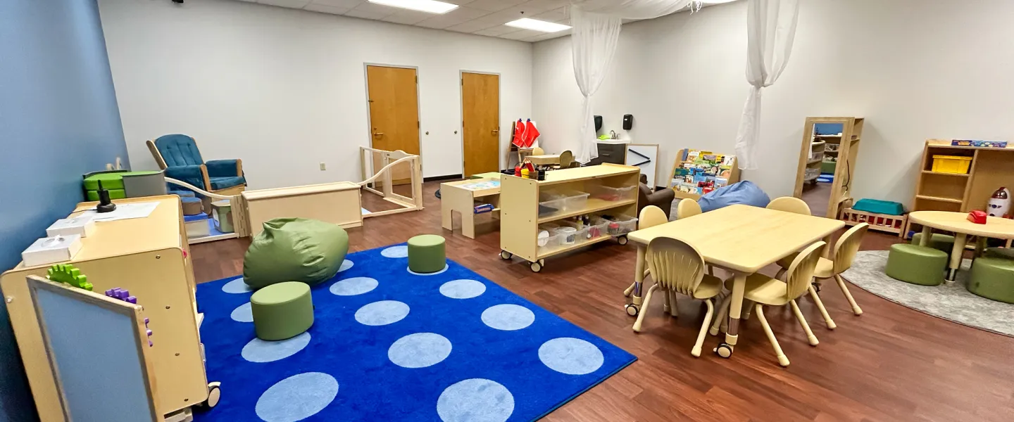 Wide view of the newly renovated YMCA Child Watch room featuring a blue accent wall, a colorful dotted rug, green bean bag chairs, and a gated infant play area.