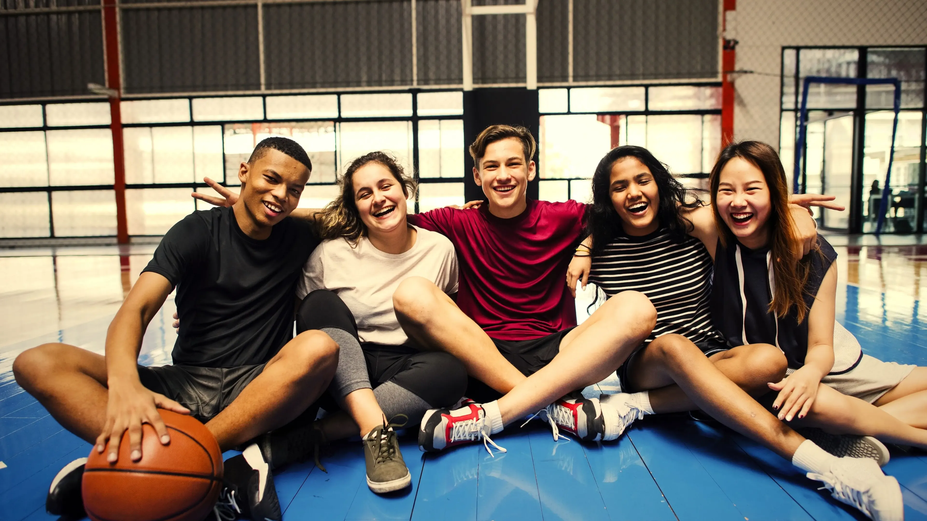 An image of a group of teenagers from the YMCA teen program linked arm-in-arm sitting on a basketball court. From left to right is an African American male, a Caucasian female and male, an African American female, and an Asian female. Everyone is smiling and the male in the middle is making peace signs on his friends shoulders.