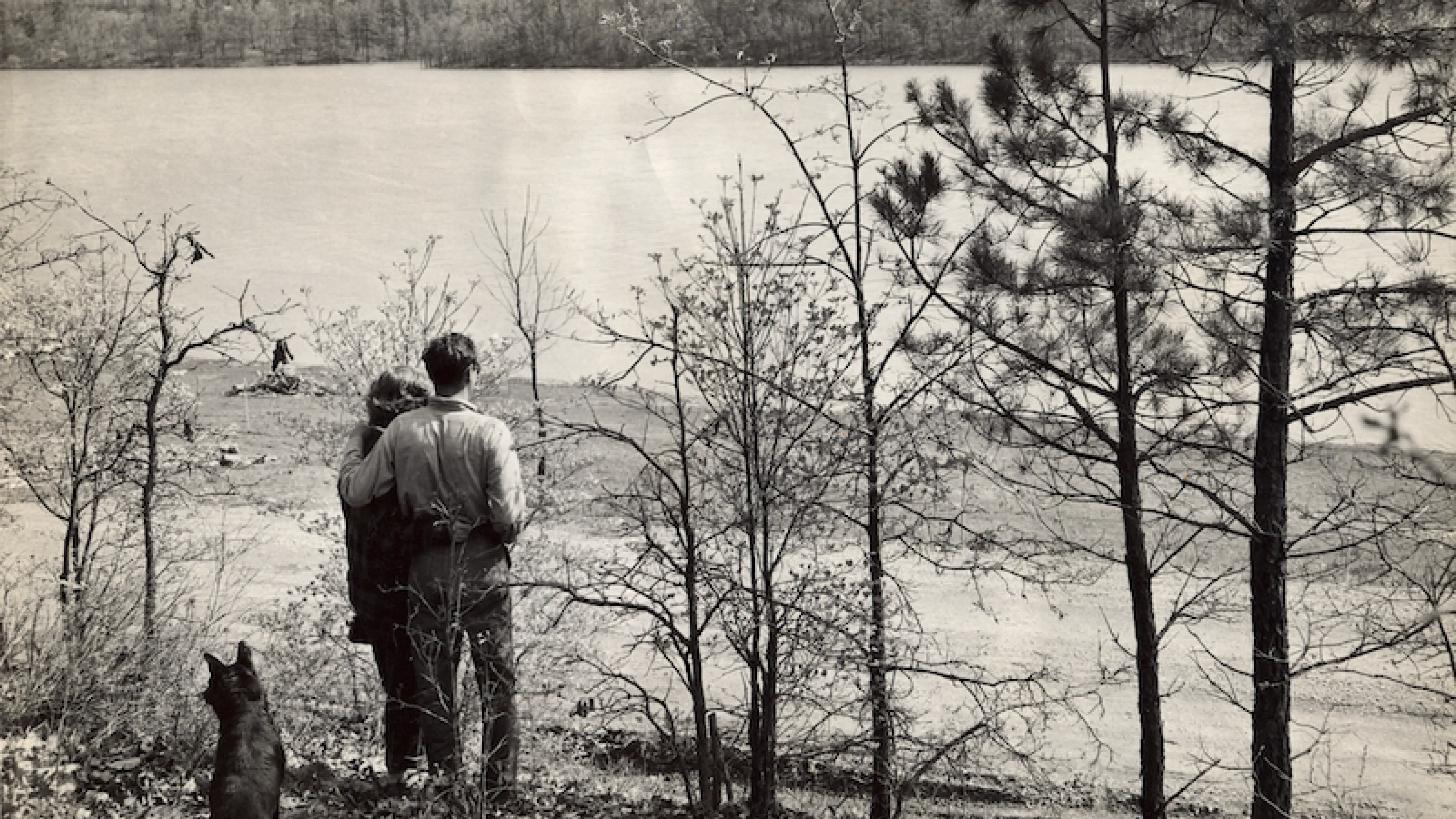 Historical photo of Sunnen Lake at YMCA Trout Lodge in Potosi, MO with a couple and their dog standing by the lake gazing out at the water and beautiful Ozark's mountain scenery