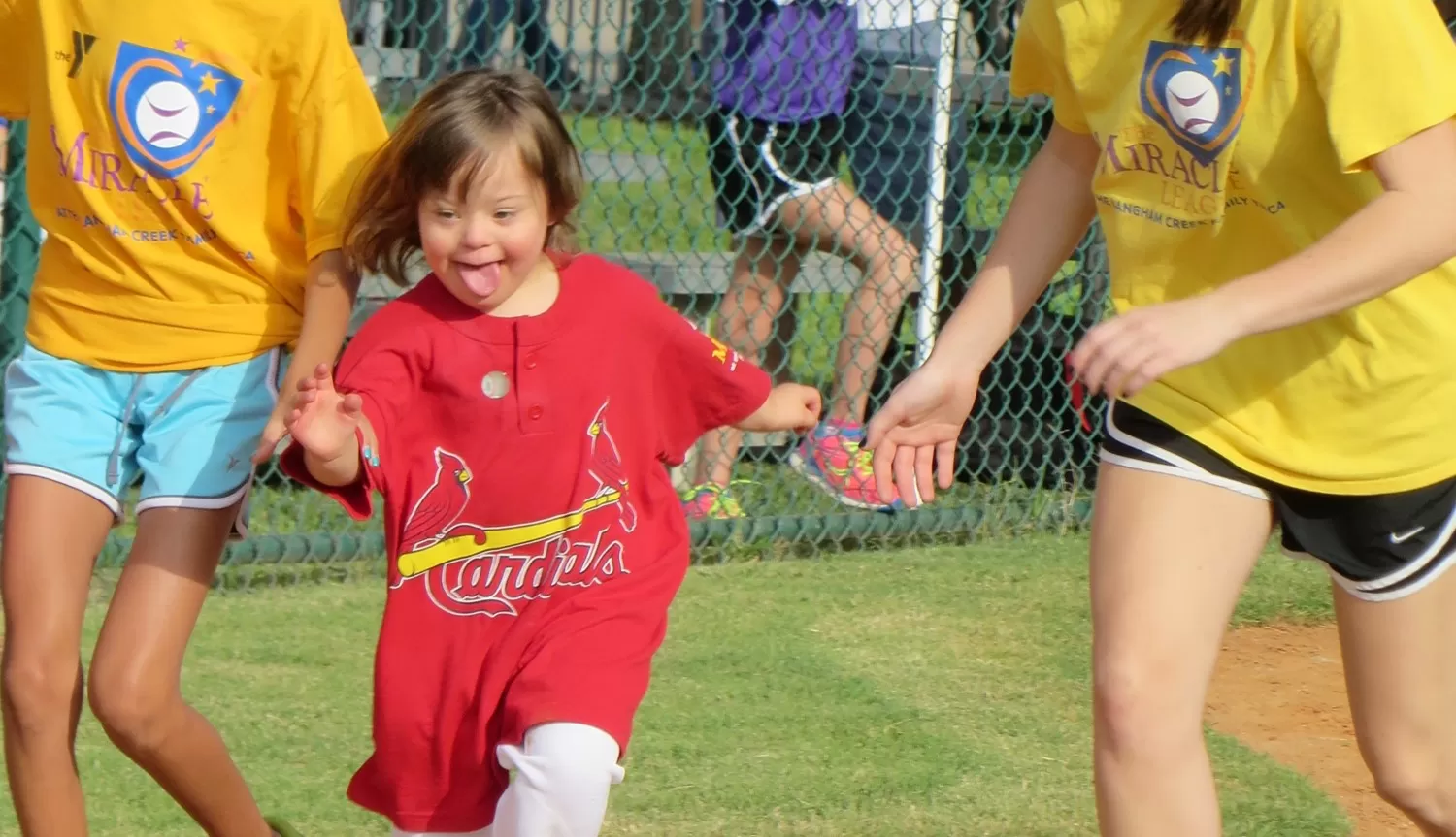 A happy young girl with Down Syndrome in the Miracle League baseball program runs to first base in a St. Louis Cardinals team jersey.