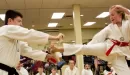 Thumbnail: A young Caucasian female kicks a board through her instructor's hands during a karate class at the YMCA. She is wearing a white karate uniform with a red belt, and her instructor is wearing the same white karate uniform except he has a black belt. A line of students waiting to test for their next belt are sitting on the ground behind them, watching.