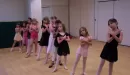 Thumbnail: A row of ten young female dancers captured learning a dance during a class at the YMCA. The girls' arms are crossed against their chest, and they are looking at themselves in the mirror whilst getting their steps down.