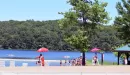 Thumbnail: An image of a waterfront that is booming with swimmers, lifeguards and boats on the lake. It was taken on a sunny summer day.