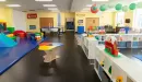 Thumbnail: Child care room with play structures, toys, separate areas for toddlers and infants, and tables. Brightly colored, fun environment.