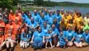 Thumbnail: A large family reunion in various t-shirt colors gathers in front of the lake