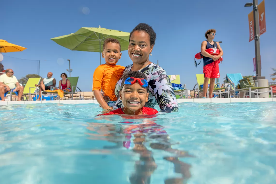 A mother and her toddler and child swim in a YMCA outdoor pool supervised by a YMCA Lifeguard