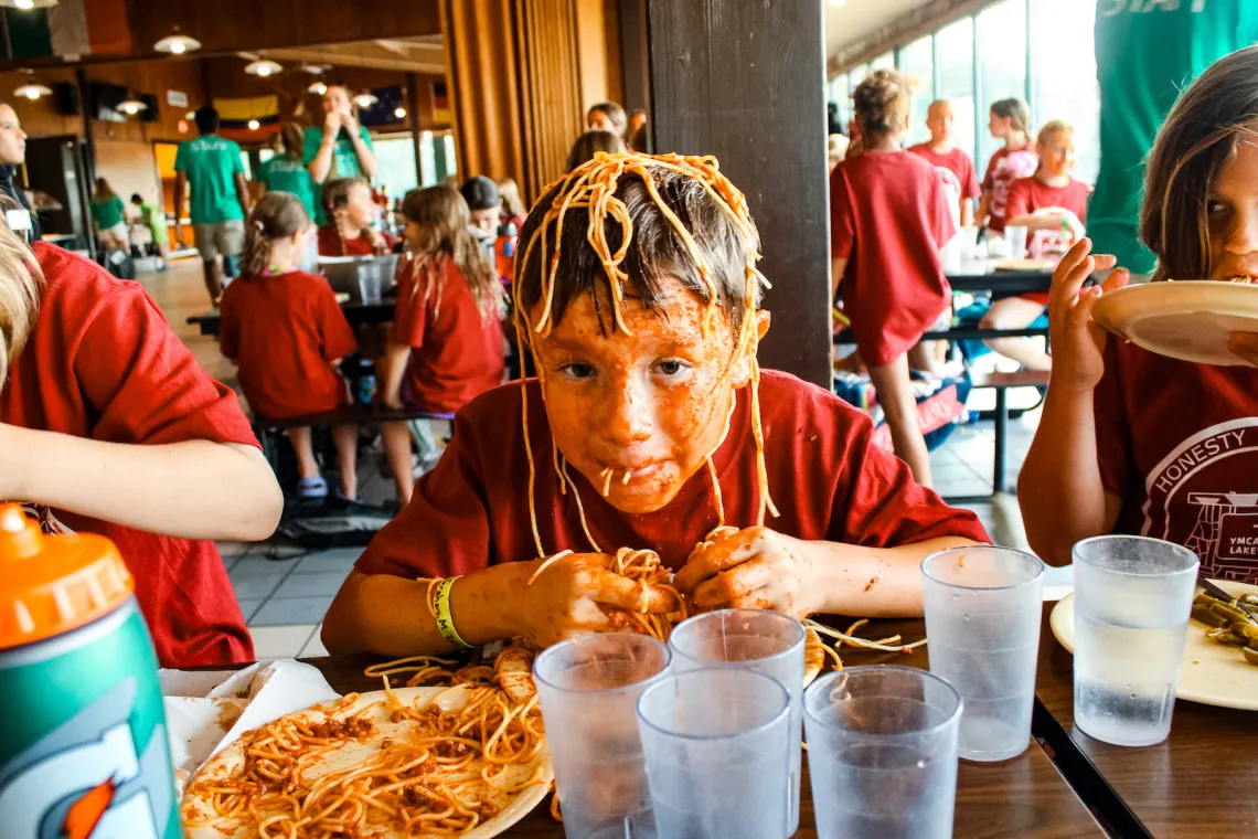 A YMCA Camp Lakewood overnight camper with his face covered in spaghetti noodles and sauce, enjoying dinner at the infamous No Utensils Needed themed dinner.