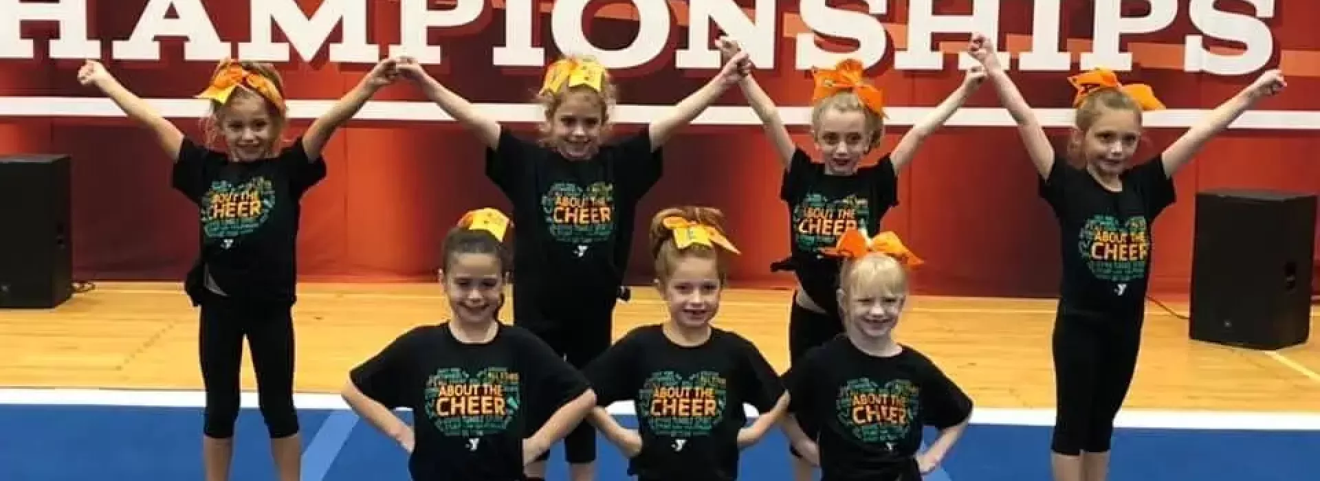An image of young, Caucasian girls competing in a cheerleading competition at the YMCA. The girls are wearing orange bows on their heads, and matching black tops paired with black pants.