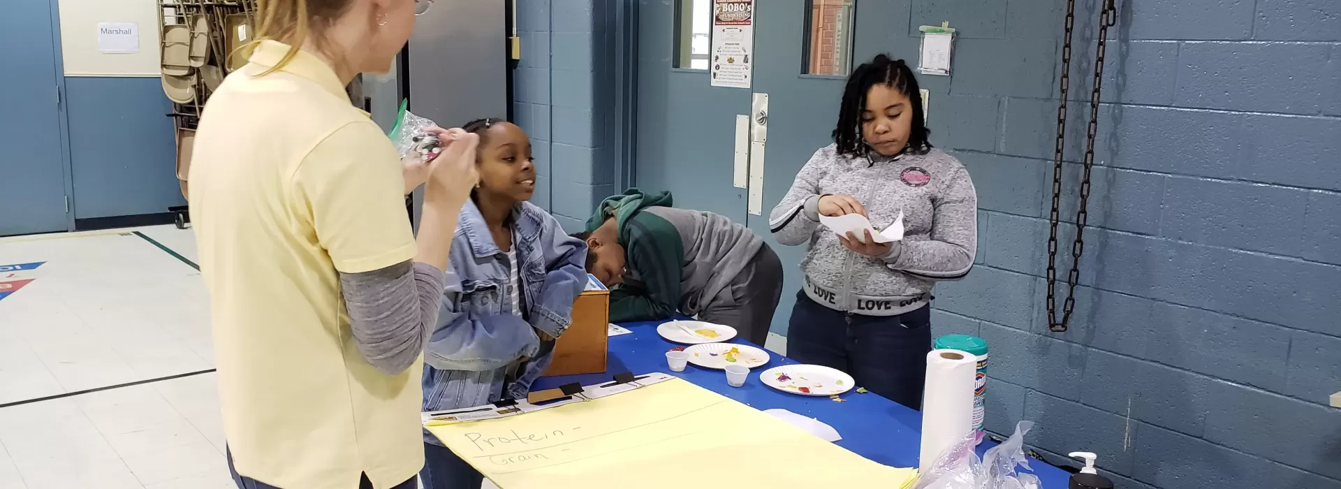 An image of a Caucasian after school leader quizzing the three African American children in her care about nutrition facts that she just taught them.