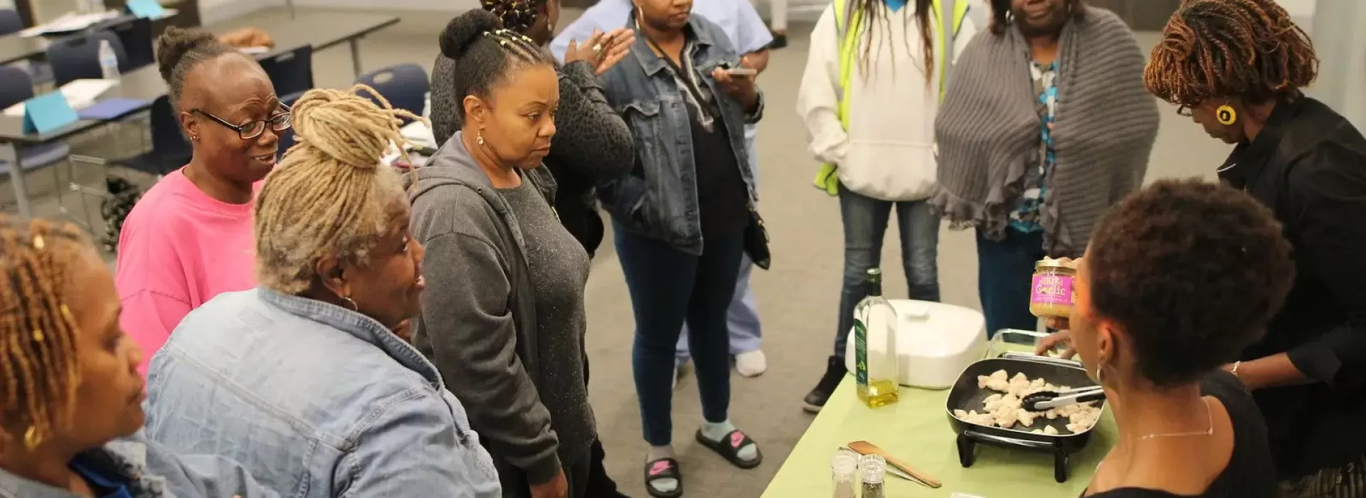 An image of a group of African American women huddled together around the class instructor's teaching them cooking skills and nutrition facts at the YMCA.