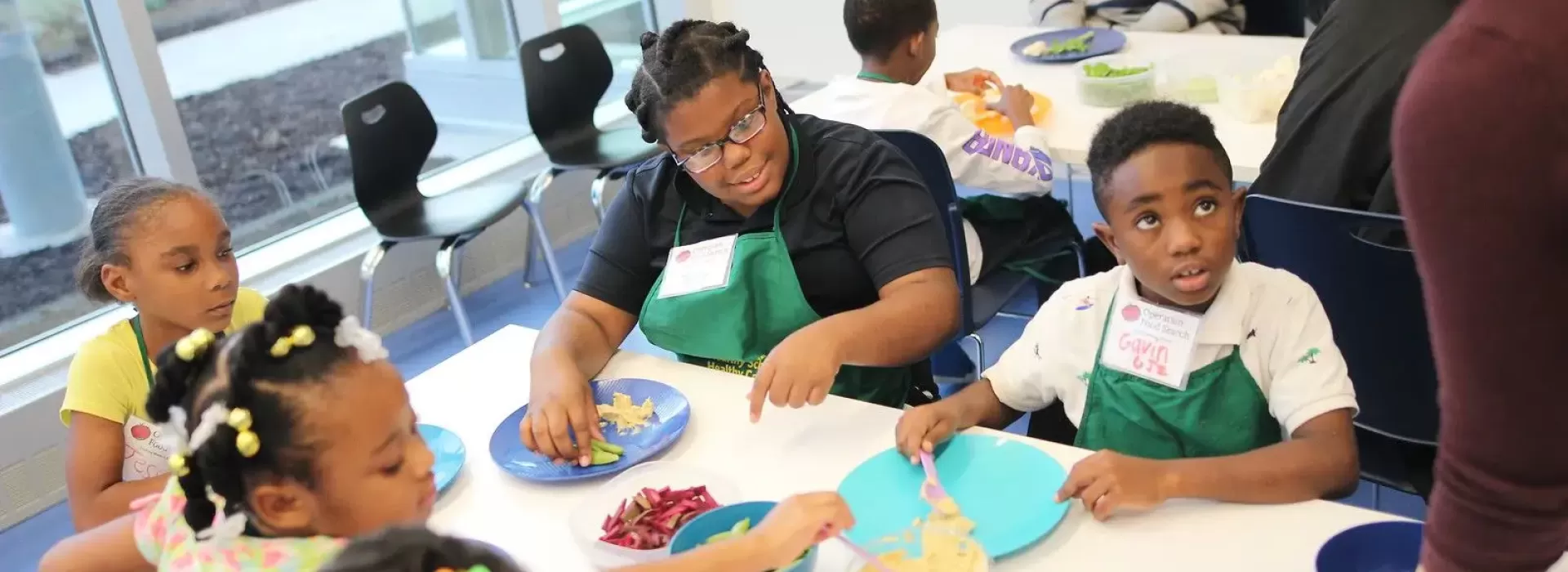 An image of a Caucasian woman wearing an apron assisting a group of young African American boys and girls during an after school program provided by the YMCA. They are cooking and learning about the importance of nutrition.