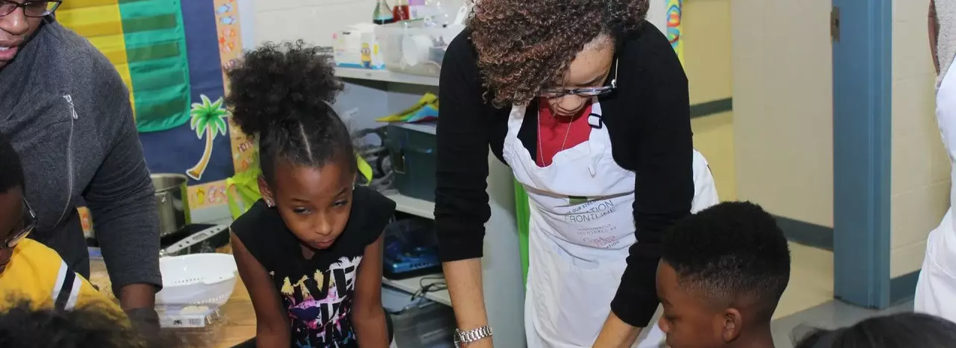 Two African American females in their middle ages helping a classroom of kids with nutrition skills. The instructor on the right is wearing a white apron over her clothes.