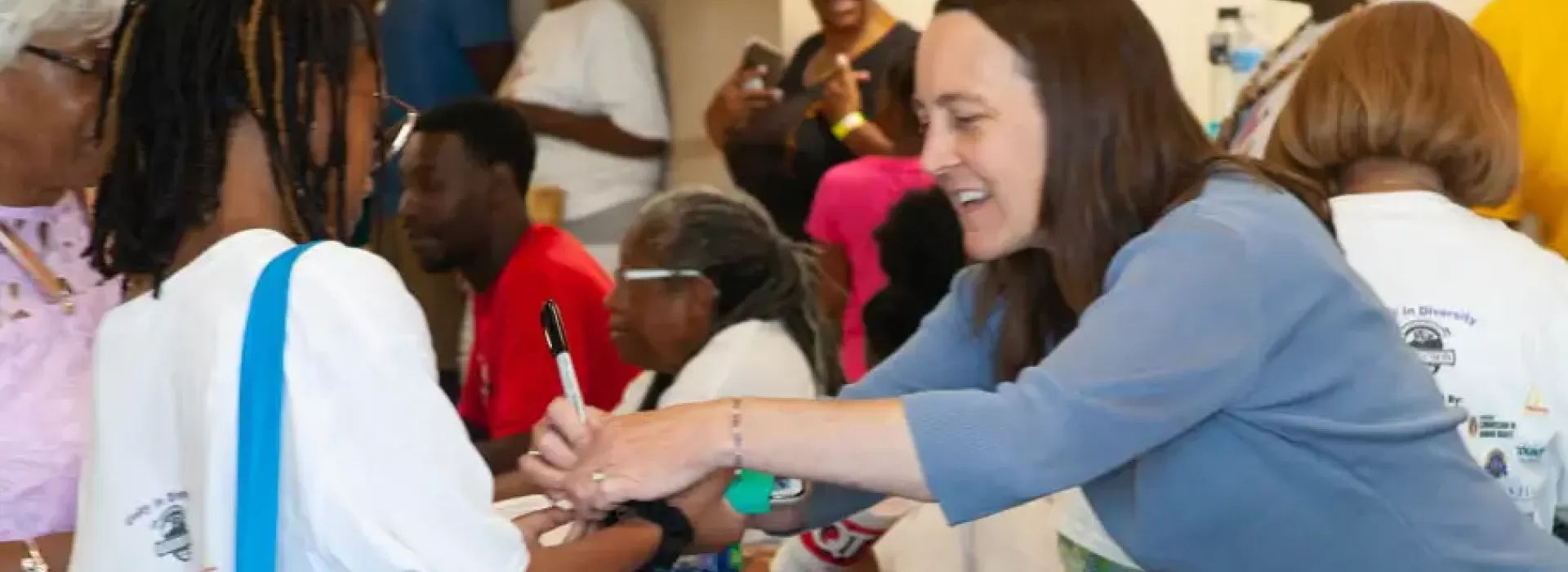 An image of a Caucasian volunteer at the YMCA's backpack give-away assisting a young African American female and her grandmother. There is a group of middle-aged African American females and males also visible in the background.